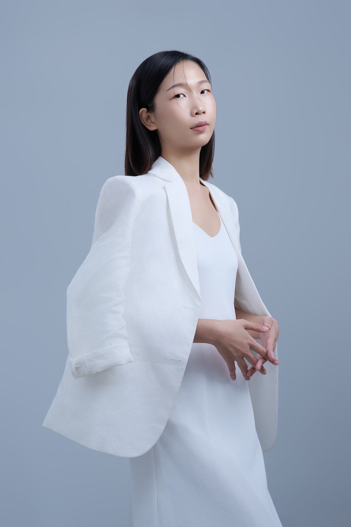 台南造型師推薦：時尚藝術風格職人肖像。彩妝造型師身穿白色披肩西裝，以優雅側身姿態展現內斂且具備高級感的專業氣場所。灰色簡約背景烘托精緻妝發，適合個人品牌形象、講師介紹與時尚媒體合作宣傳。