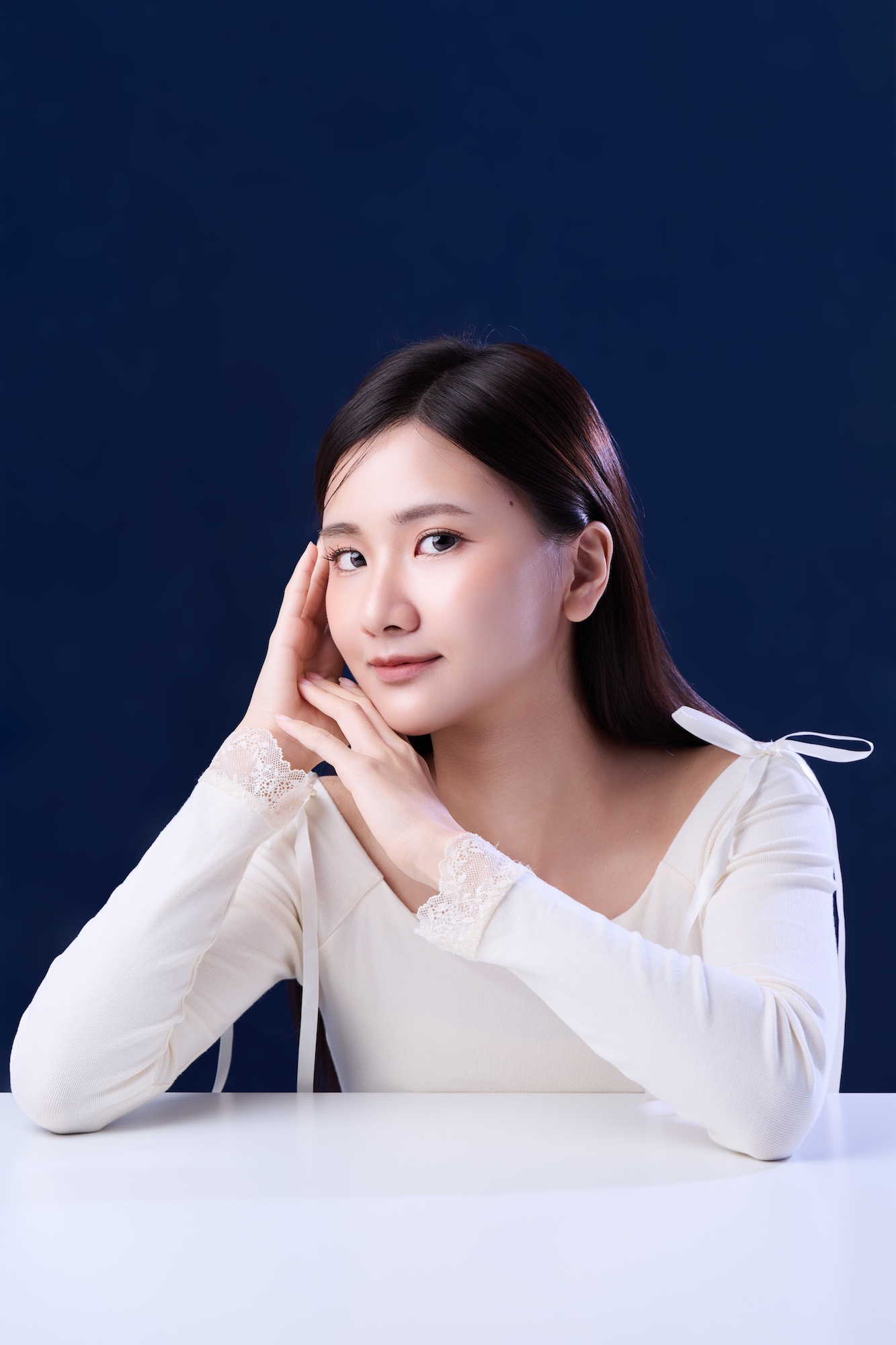 台南形象照首選：熱銷款韓系精緻肖像。長髮女性身穿白色蕾絲袖上衣，以優雅雙手托腮姿勢展現溫柔氣質。深藍背景強烈襯托通透膚質，適合個人品牌、精緻履歷與社群頭像。
